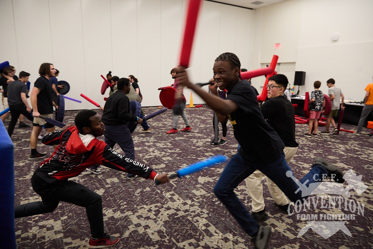 A full convention room of fighters plays a fast-paced foam battle with red and blue padded weapons.