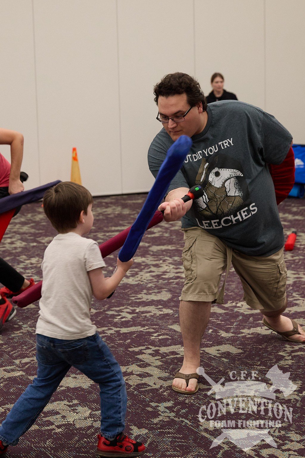 An adult and a child safely practice foam fighting together using padded weapons at a convention event.