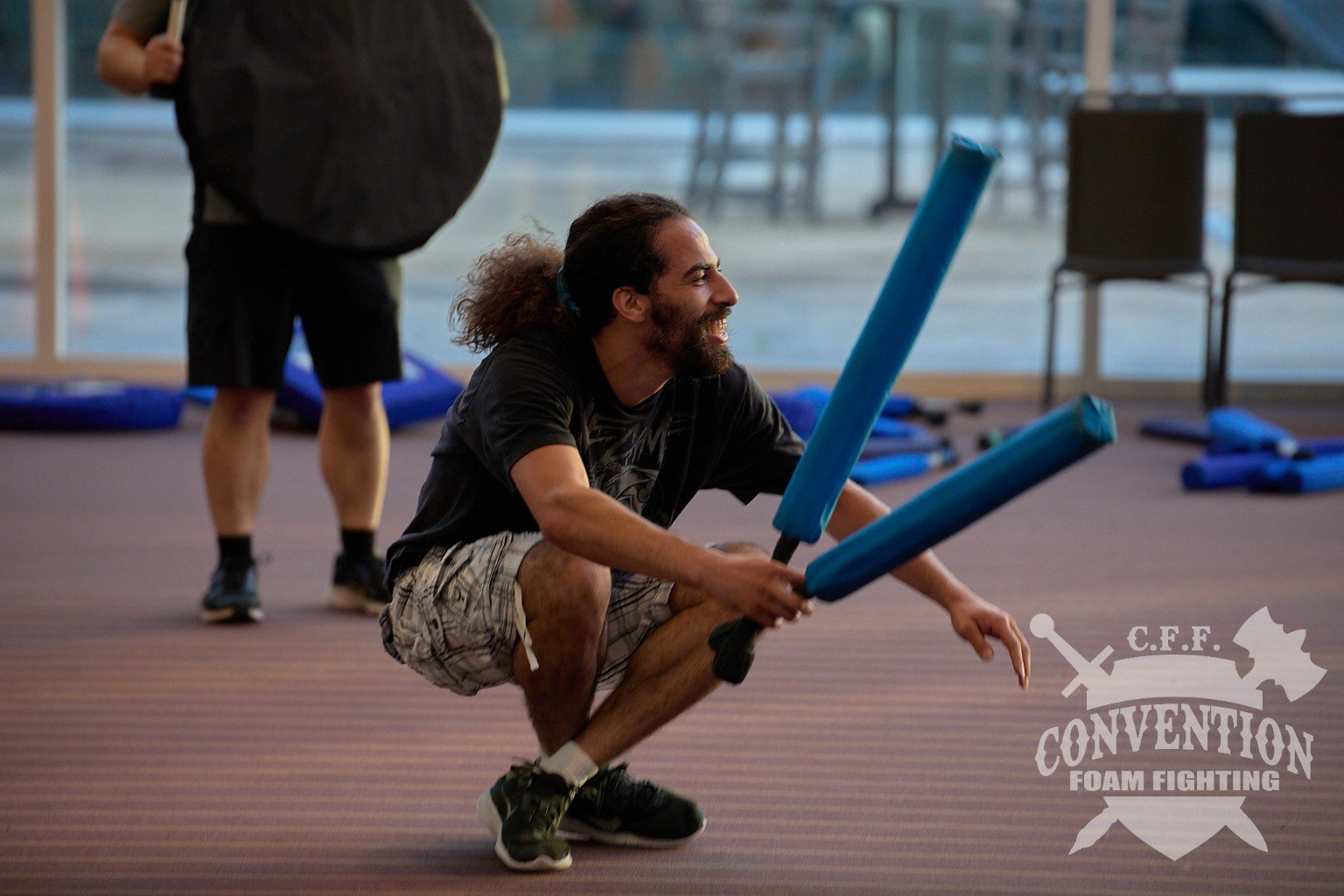A smiling participant crouches with two blue foam weapons while other players and shields sit in the background.